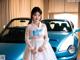 A woman in a white dress standing next to a blue car.